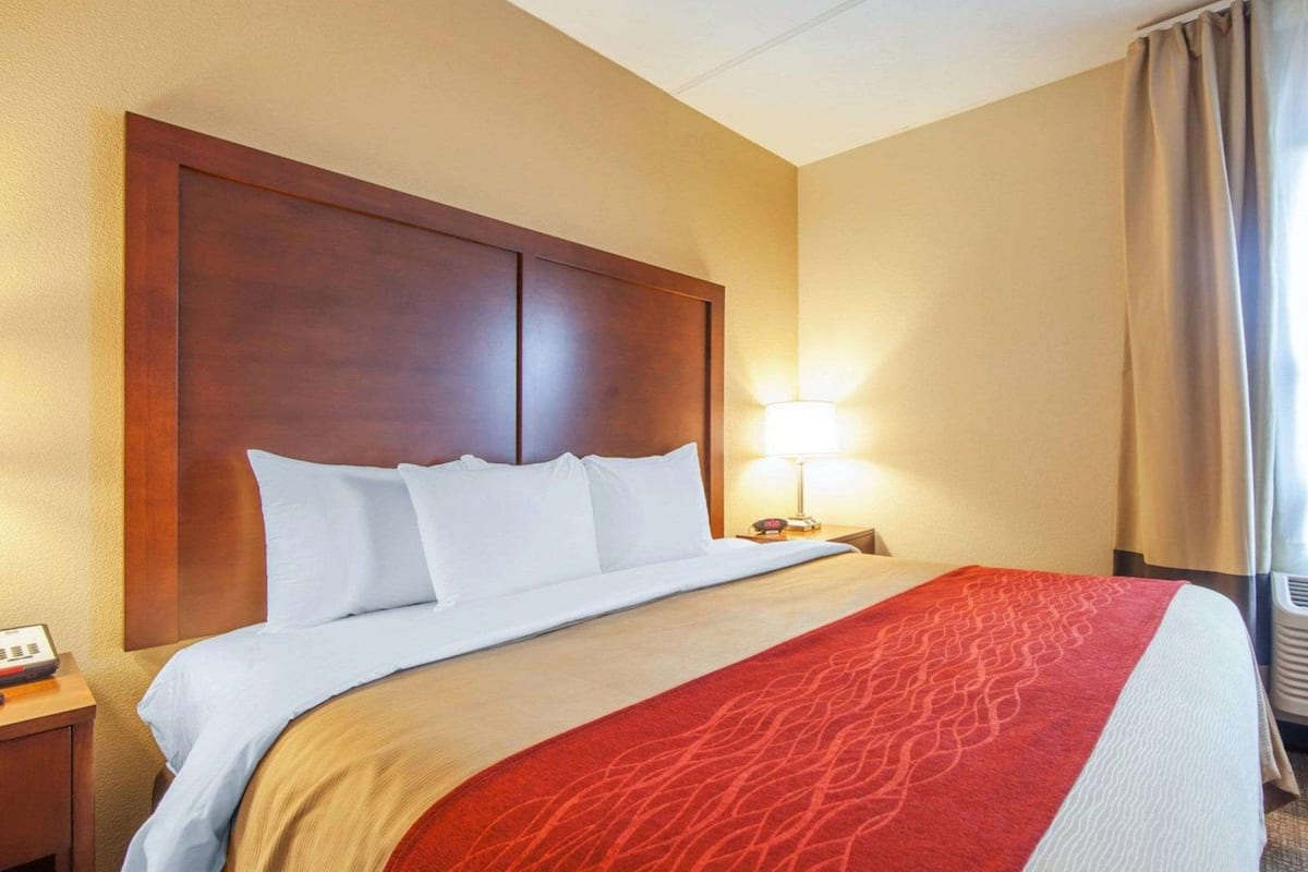 A hotel room with a wooden headboard, a bed with white bedding, a red patterned throw, a bedside lamp, and a window with curtains.