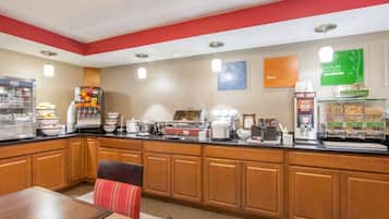 Una barra de desayuno con diversas opciones de comida y bebida, una zona de estar con mesa y sillas, y una pared decorada con carteles motivacionales.