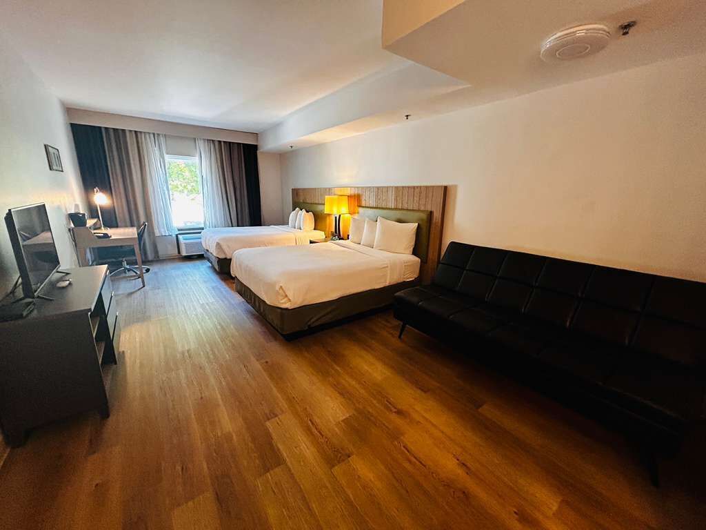 A hotel room with two beds, a desk with a computer, a chair, a black sofa, and a window with curtains.