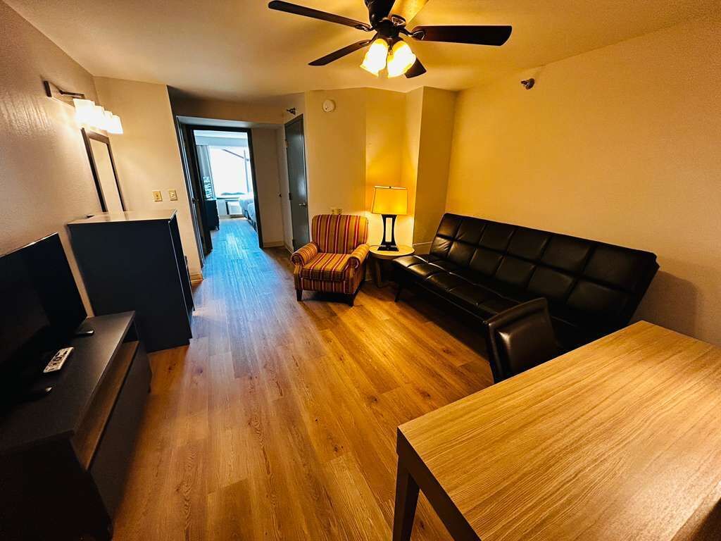 Una sala de estar con piso de madera, un sofá negro, un sillón a rayas, una mesa, un ventilador de techo y una lámpara.