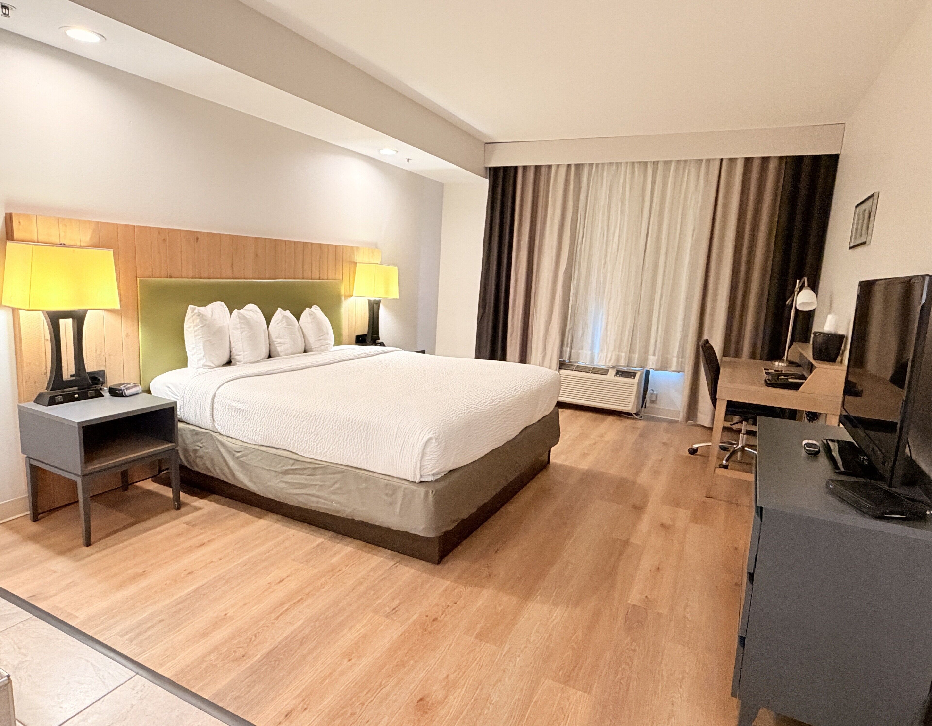 A hotel room with a bed, bedside tables, a desk, a chair, a television, and a window with curtains.