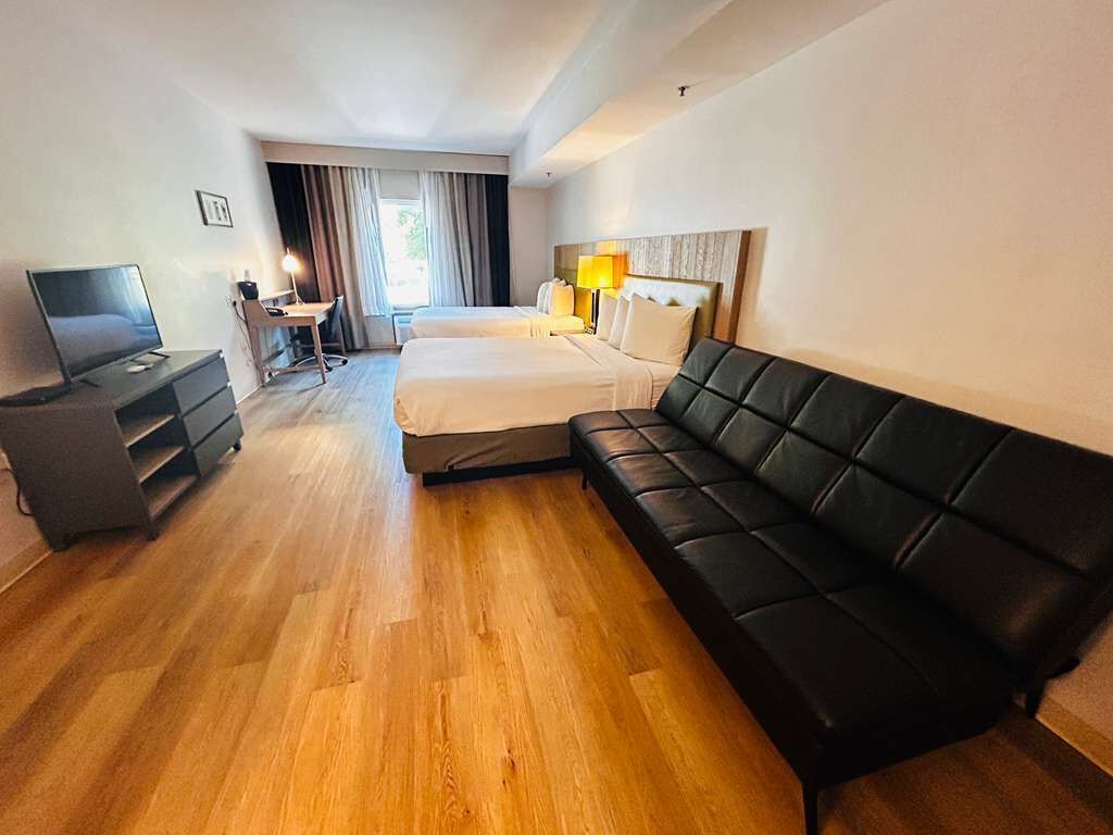 Una habitación de hotel con piso de madera, un sofá de cuero negro, una cama con ropa blanca, un escritorio con una silla, un televisor y una lámpara.