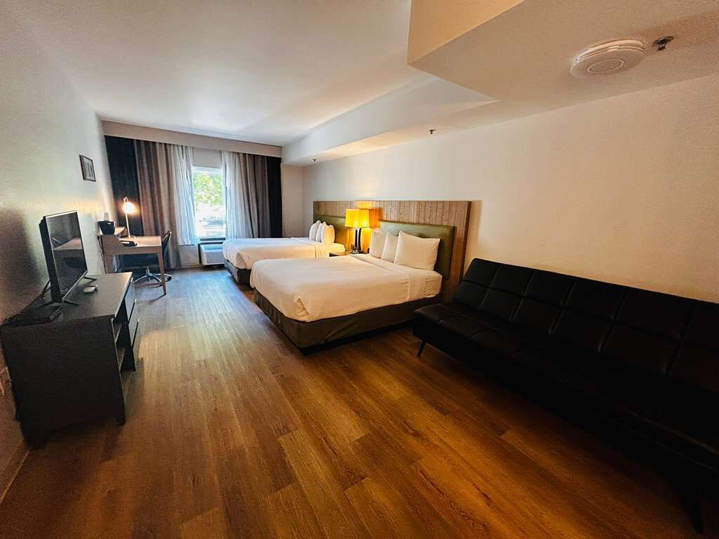 Une chambre d’hôtel avec deux lits, un bureau, une chaise, un canapé noir et une fenêtre avec des rideaux.