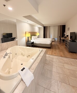 A modern hotel room with a large bathtub, a bed with a wooden headboard, a desk with a chair, and a television. - Radiant Inn & Suites (Murfreesboro)