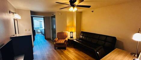 A room with a wooden floor, a black sofa, a striped armchair, a small table, a ceiling fan, and a lamp.