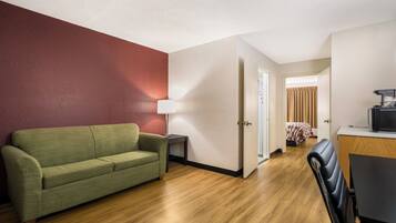 A room with a green sofa, a lamp, a wooden floor, a bed with a checkered blanket, and a kitchenette area.