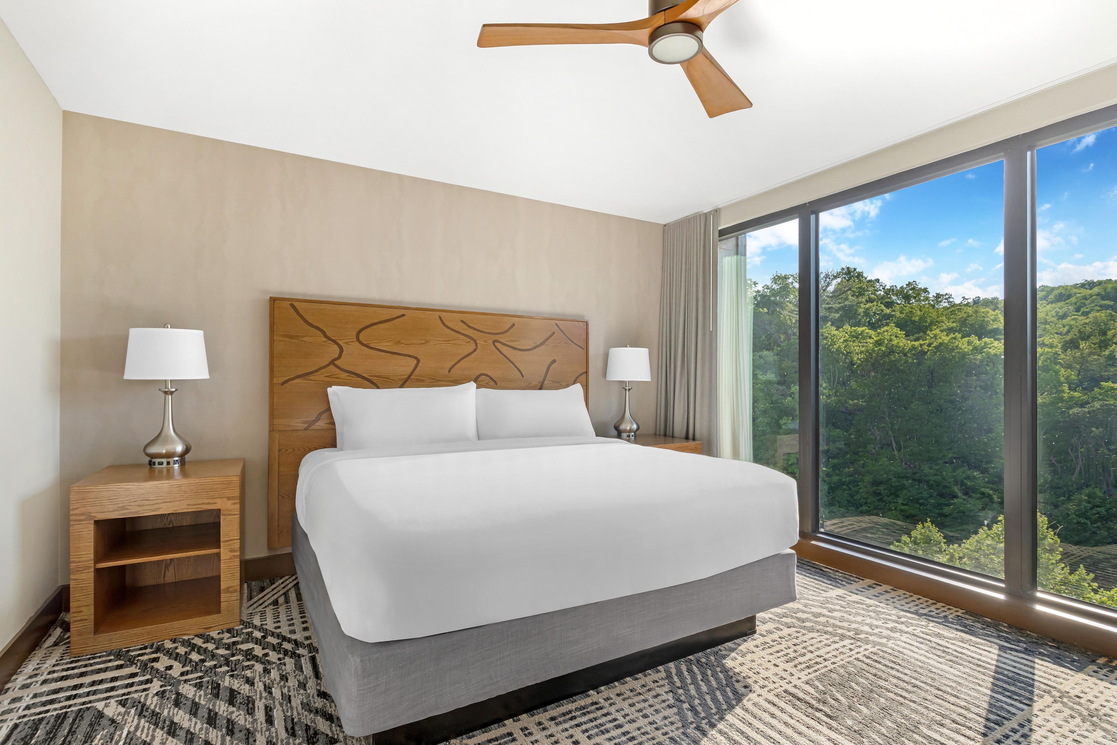 Habitación de hotel con una cama grande, dos mesitas de noche, un ventilador de techo y un ventanal con vistas a árboles.