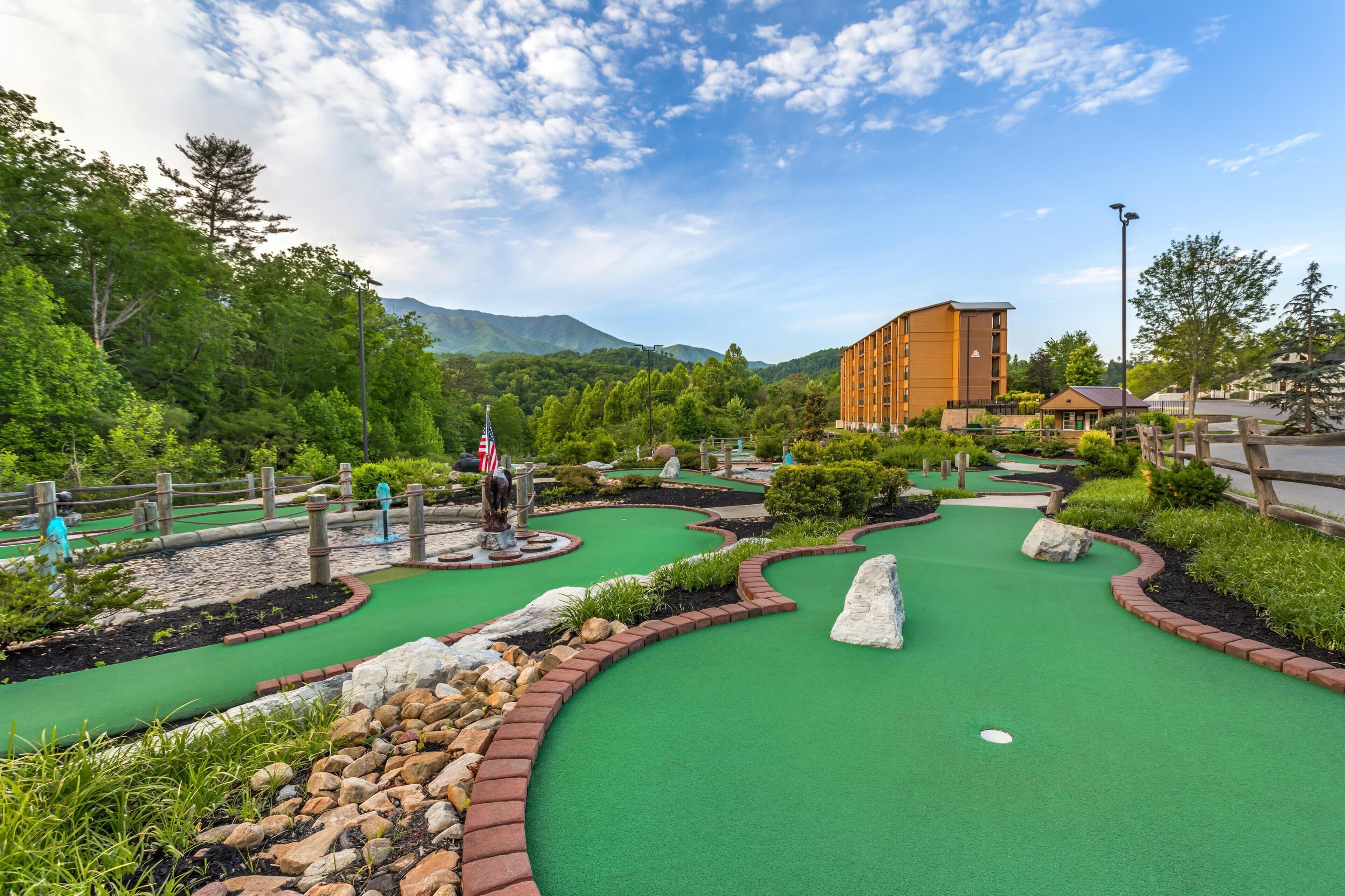 Un parcours de mini-golf doté d’un green, entouré d’espaces aménagés avec des rochers et des arbustes, et avec un bâtiment visible au fond.