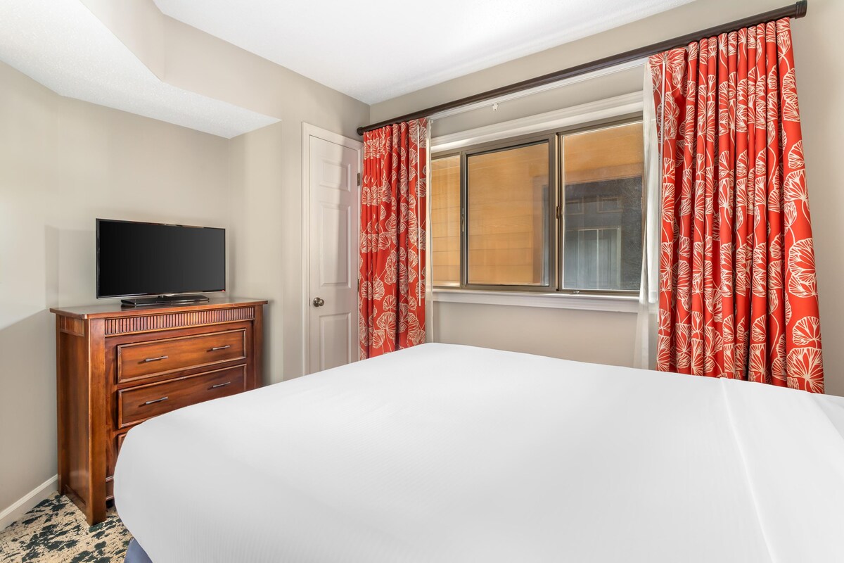 A hotel room with a bed, a wooden dresser, a television, and red curtains with a leaf pattern.