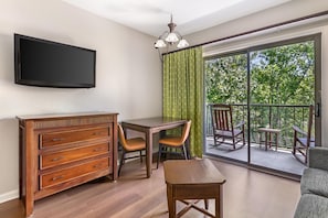 Una habitación con una televisión de pantalla plana montada en la pared, un aparador de madera, una mesa de comedor con sillas y un balcón con vistas a la vegetación.