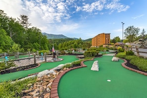 Un campo de mini golf con una superficie de césped verde, rodeado de zonas ajardinadas con rocas y arbustos, y un edificio al fondo.