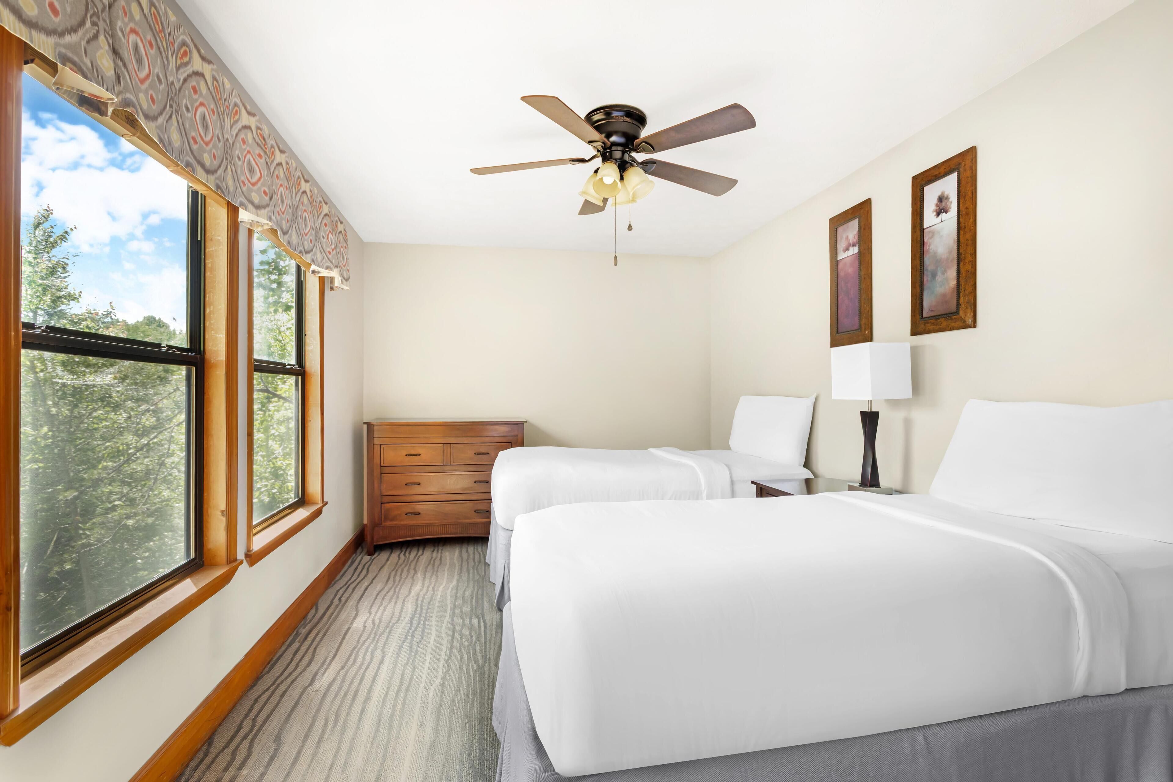 A hotel room with two beds, a ceiling fan, a dresser, and a window with a view of trees.