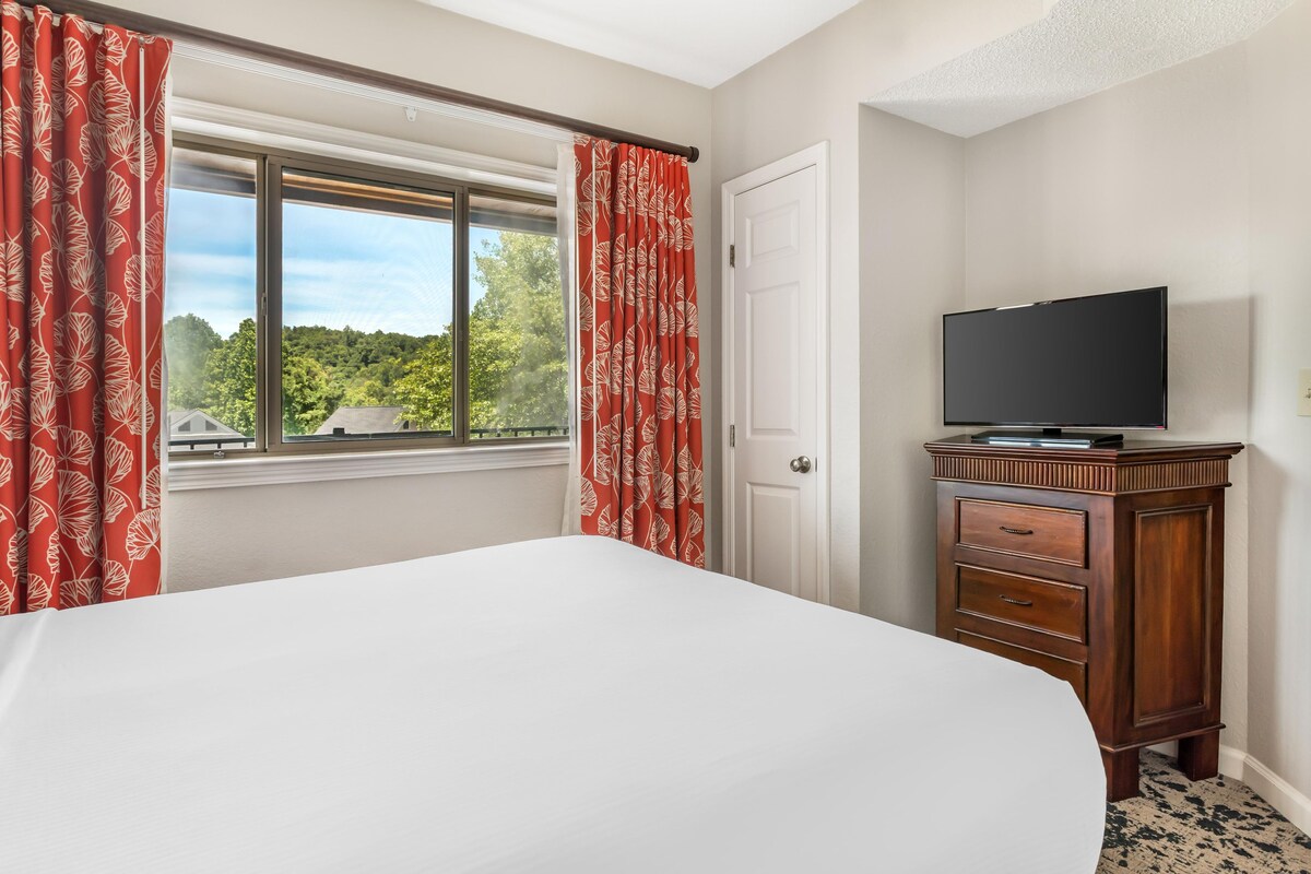 A hotel room with a bed, a television on a wooden cabinet, and a window with red curtains.