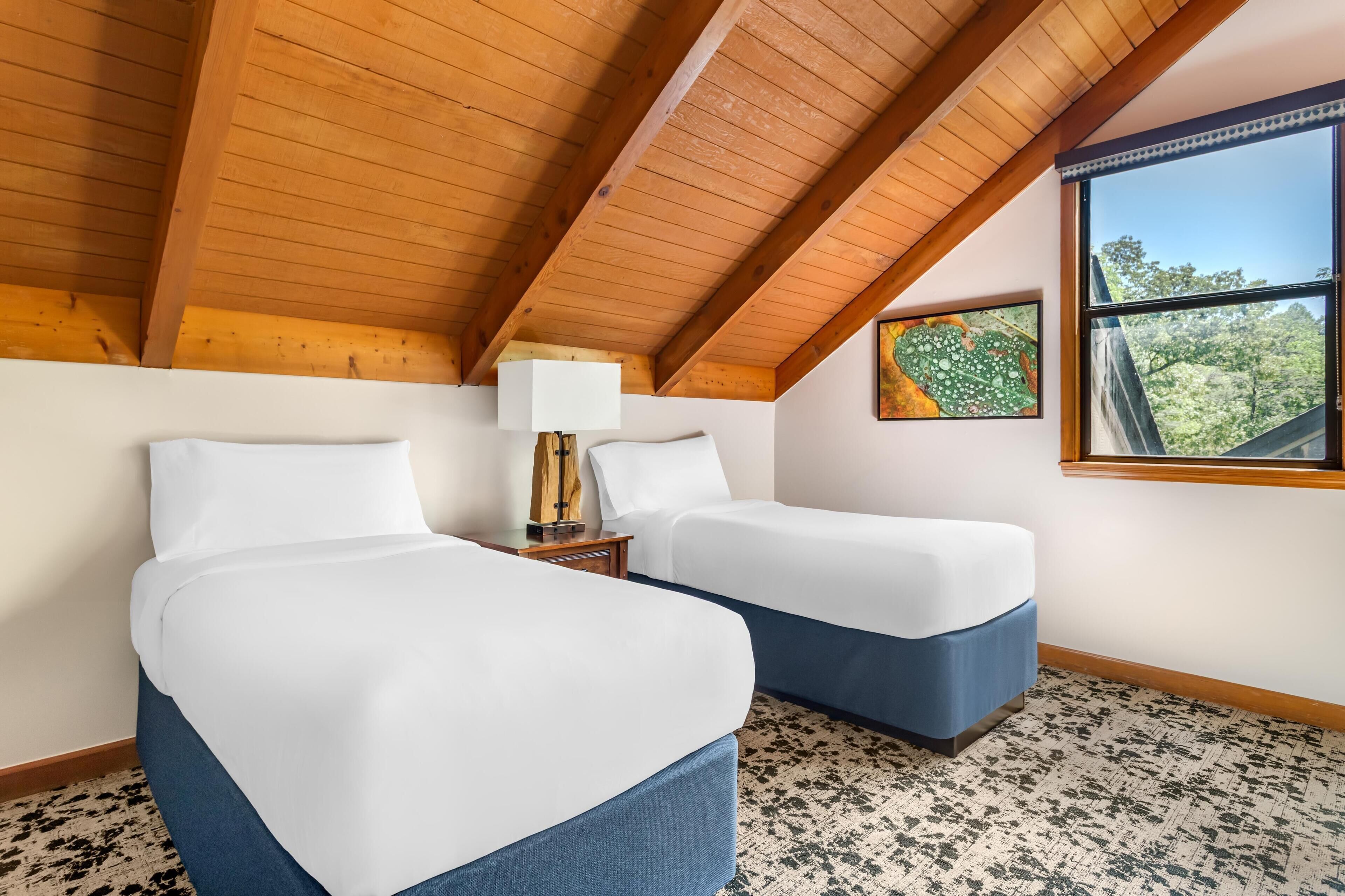 A room with two beds, a nightstand, a lamp, and a window with a view of trees.