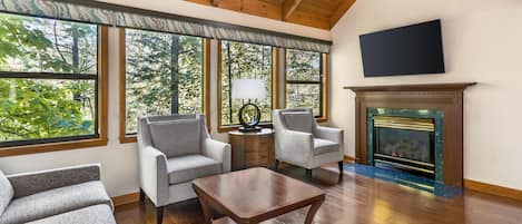 A spacious living room with a fireplace, two grey armchairs, a coffee table, and large windows offering a view of trees.