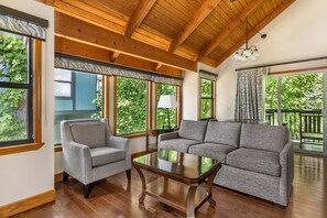 Una habitación con techo de madera, ventanas amplias con cortinas, un sofá gris, un sillón gris y una mesa de centro de madera.