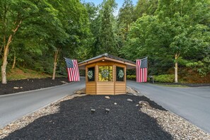Una pequeña cabaña de madera con dos banderas estadounidenses a cada lado, rodeada por un camino de grava y árboles.