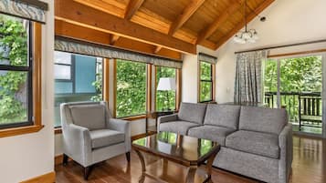 A room with a wooden ceiling, large windows with curtains, a grey sofa, a grey armchair, and a wooden coffee table.