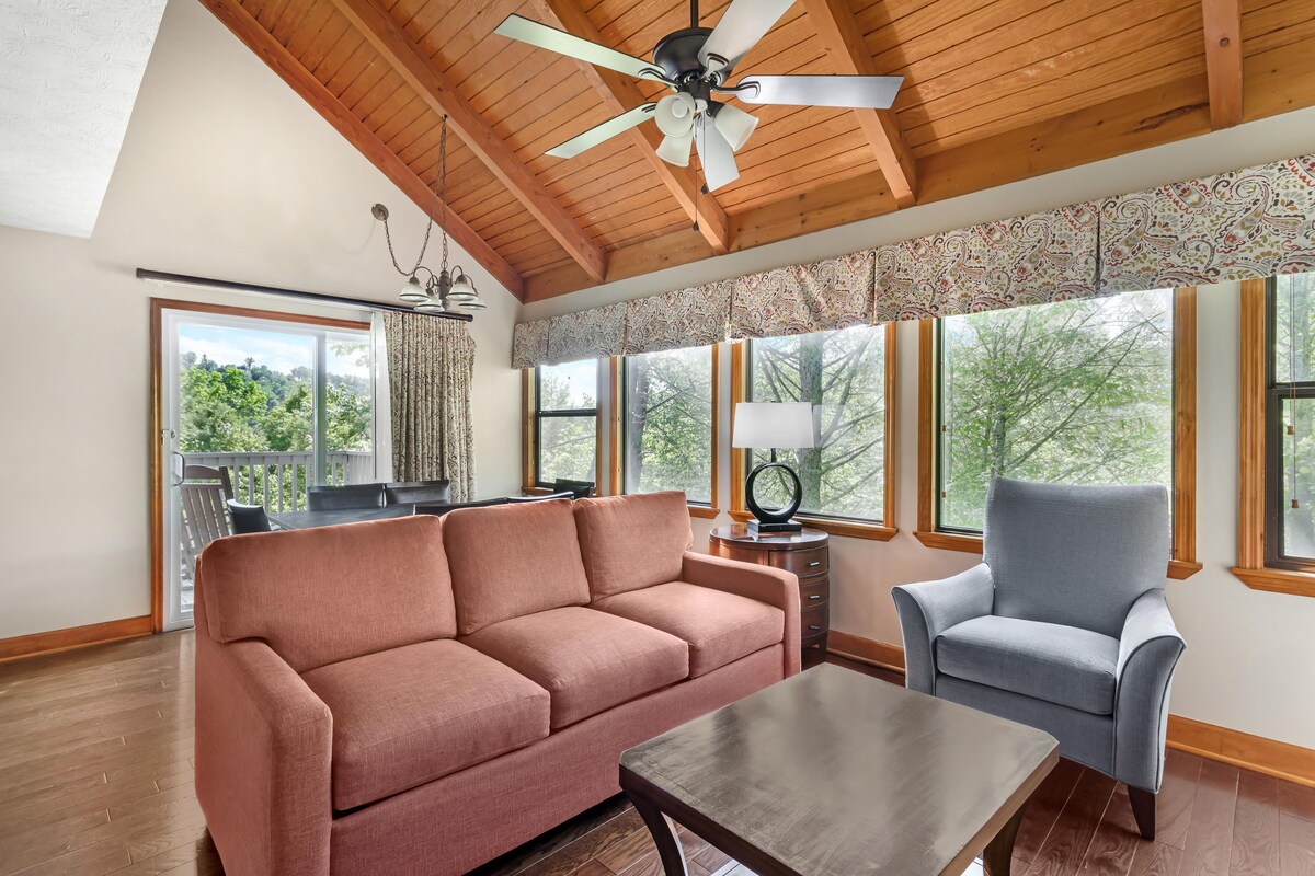 A room with a wooden ceiling, a ceiling fan, a sofa, a chair, a table, and large windows with a view of trees.