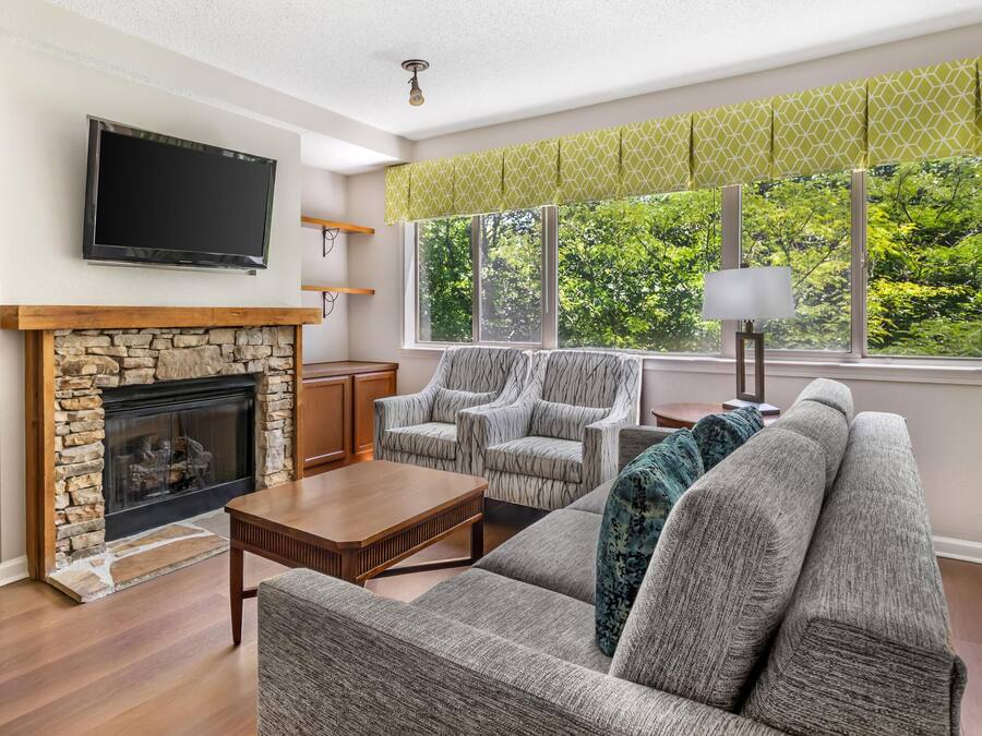 A living room with a stone fireplace, a flat-screen TV, a sofa, armchairs, and a view of greenery through large windows.