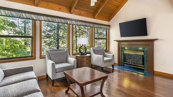 A spacious living room with a fireplace, two grey armchairs, a coffee table, and large windows offering a view of trees.