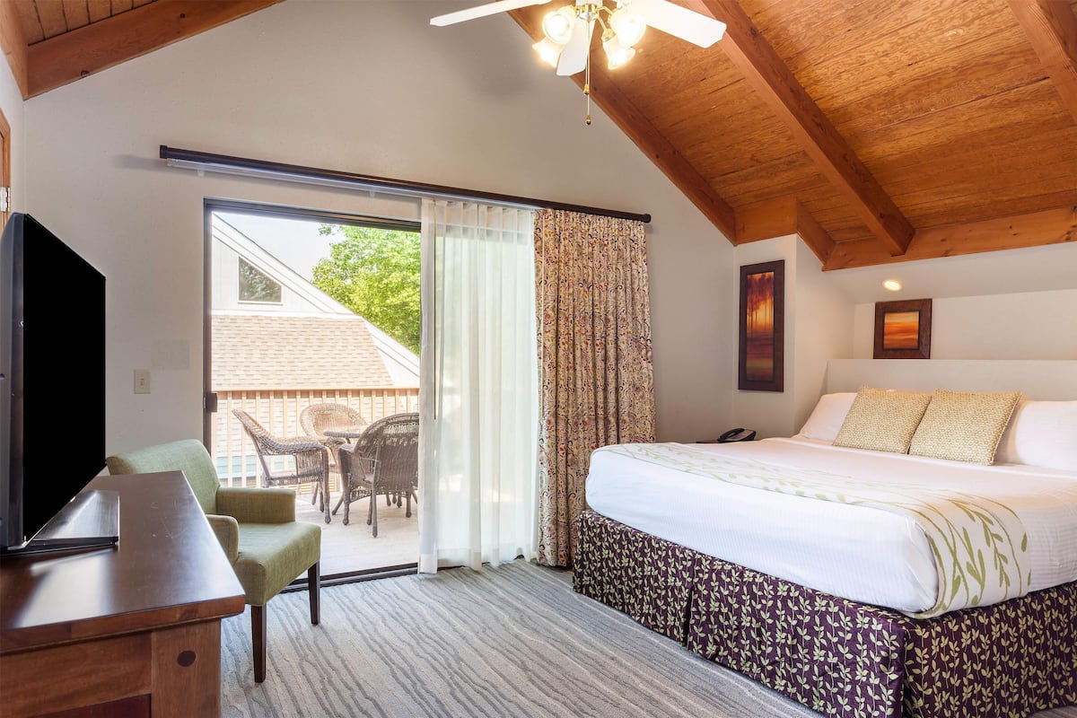 Une chambre d’hôtel équipée d’un lit, d’un bureau, d’une télévision, d’un ventilateur de plafond et d’une porte-fenêtre coulissante donnant sur un espace extérieur aménagé avec des chaises.