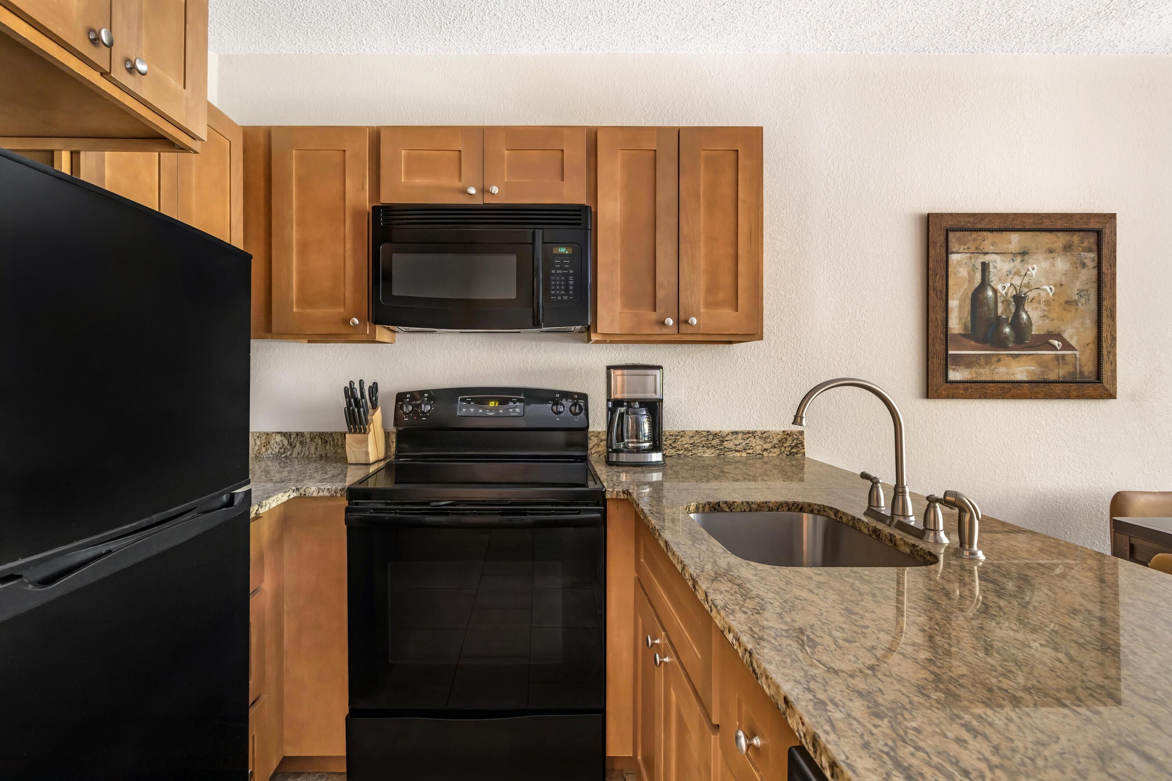 A kitchen with wooden cabinets, a black refrigerator, a microwave, a stove, a coffee maker, and a sink.