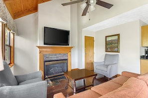 Una sala de estar con chimenea, televisor montado encima, un sillón gris, sofá y un cuadro en la pared.
