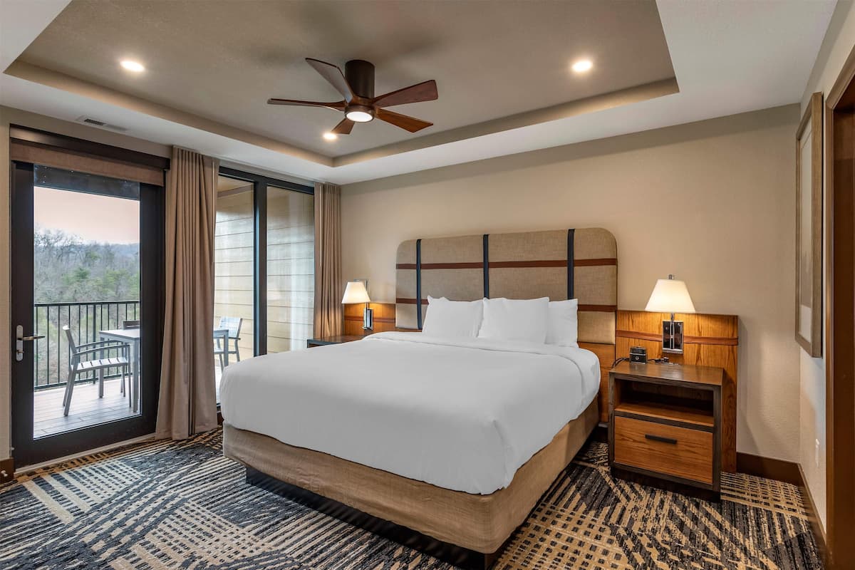 Une chambre d’hôtel avec un grand lit, deux tables de chevet avec des lampes, un ventilateur de plafond et une vue sur un espace extérieur aménagé avec des tables, accessible par une porte-fenêtre coulissante.