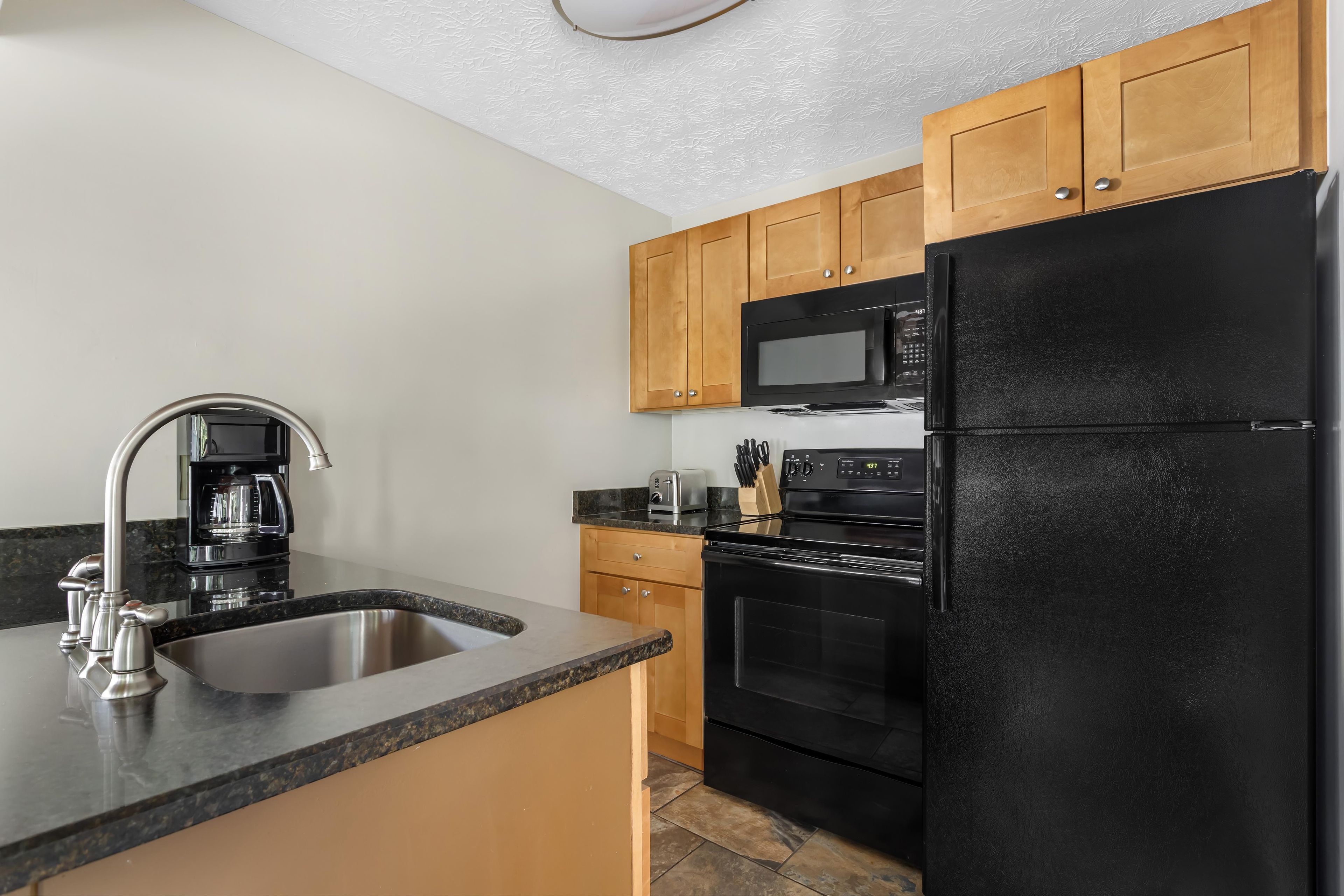 A kitchen with a stainless steel sink, a black refrigerator, wooden cabinets, a microwave, and a coffee maker.
