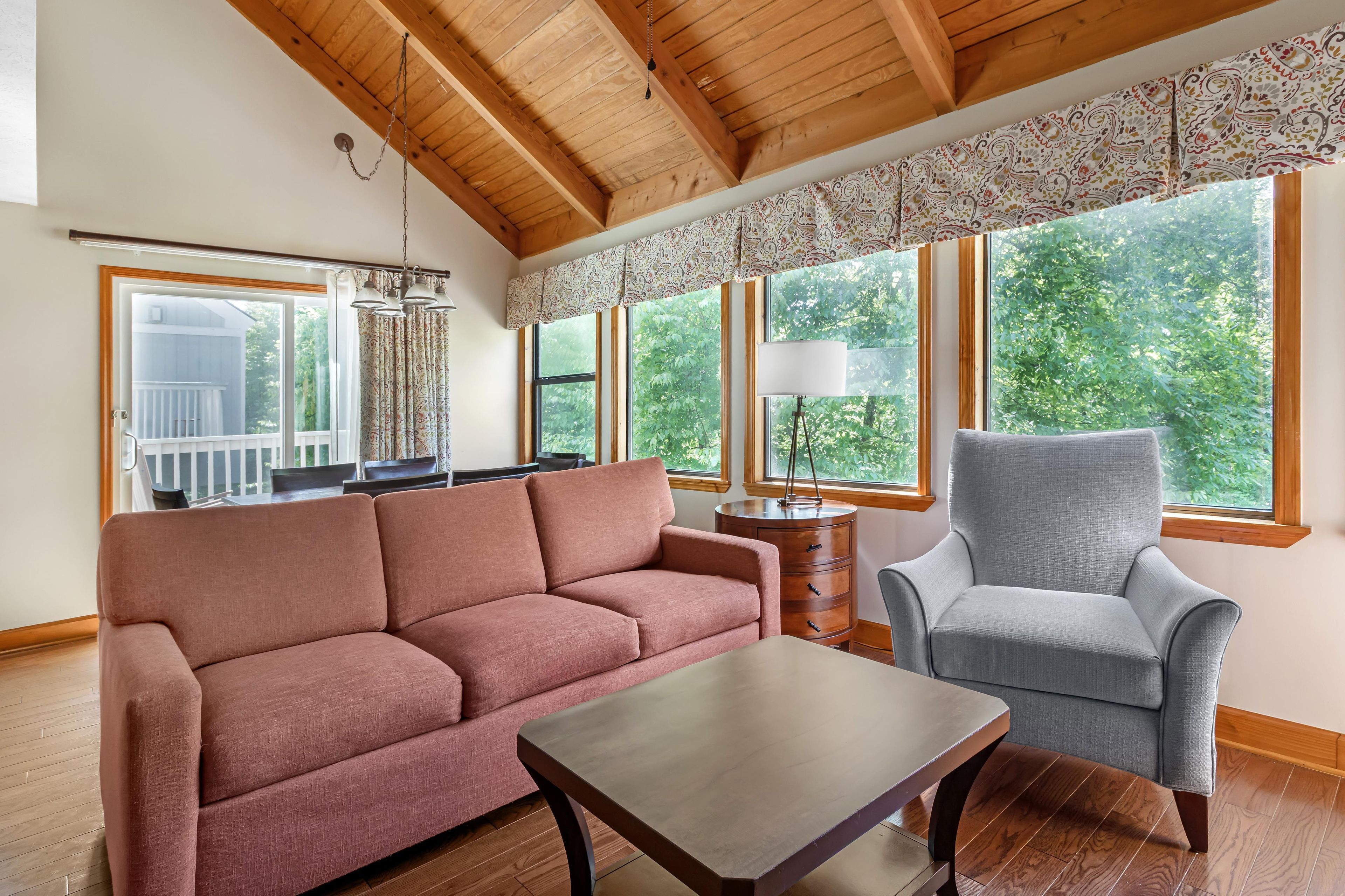 A living room with a wooden ceiling, a pink sofa, a grey armchair, a wooden coffee table, and large windows with a view of greenery.