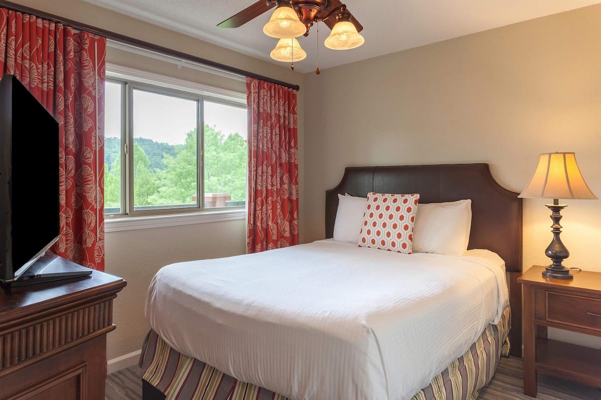 Une chambre d’hôtel avec un grand lit, un téléviseur posé sur une commode en bois, une fenêtre avec des rideaux rouges et un ventilateur de plafond.