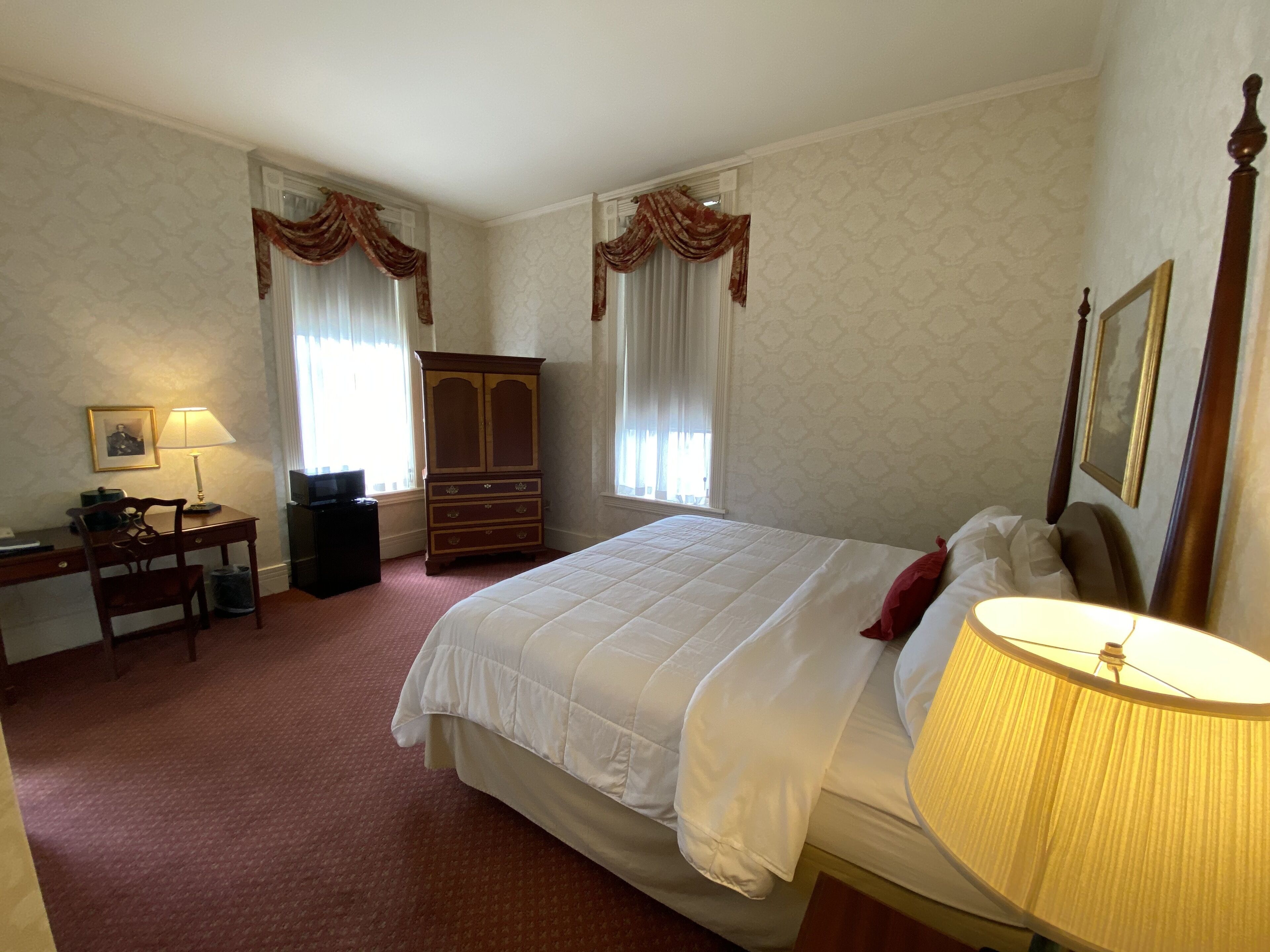 Quarto de hotel com cama, escrivaninha, cadeira, guarda-roupa e abajur.