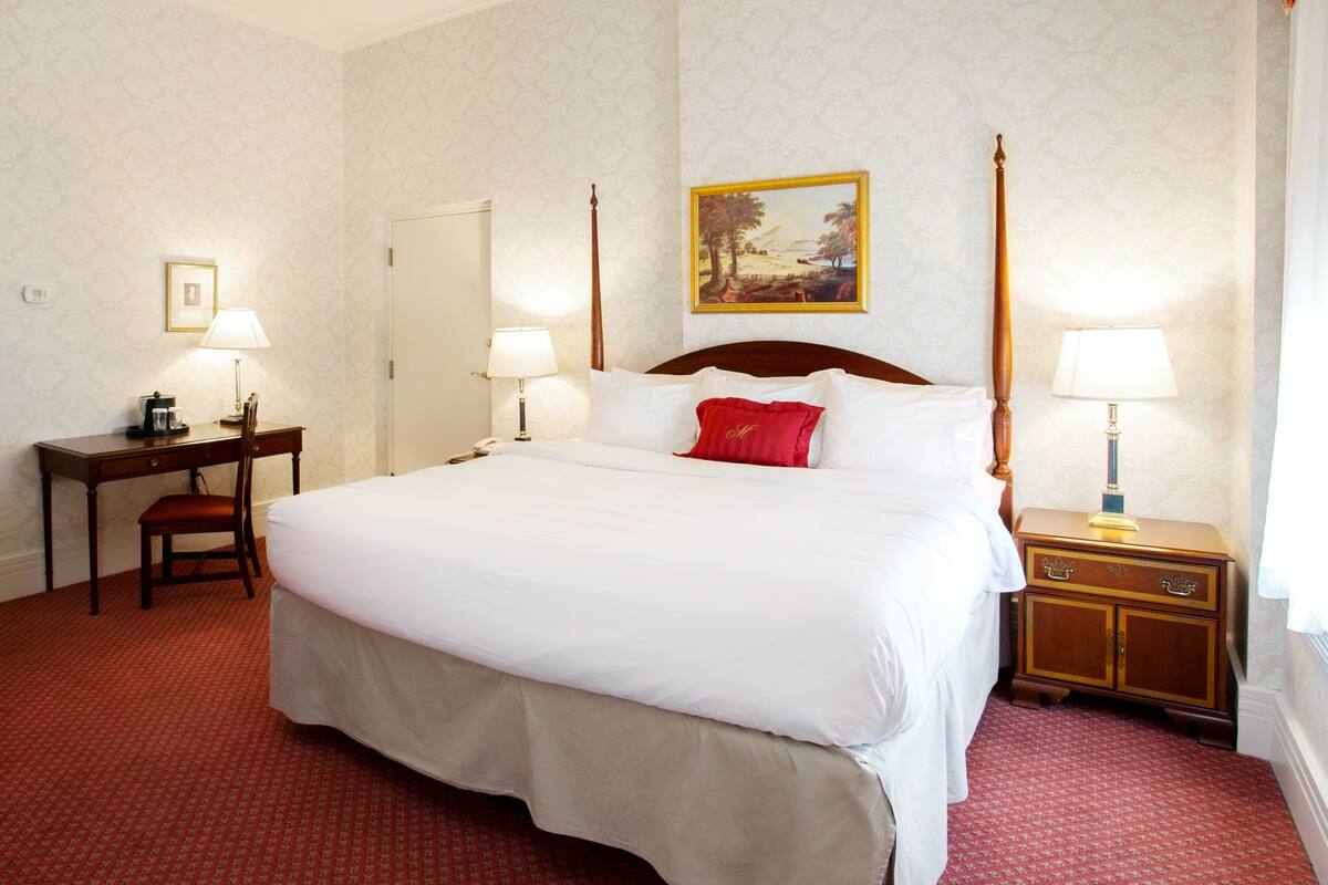 A hotel room with a large bed, two bedside tables with lamps, a desk with a chair, and a painting on the wall.