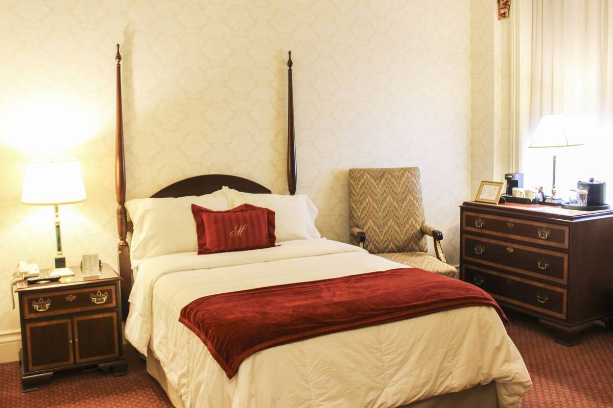 A four-poster bed with a red throw blanket, a nightstand with a lamp, a chair, and a dresser with drawers.