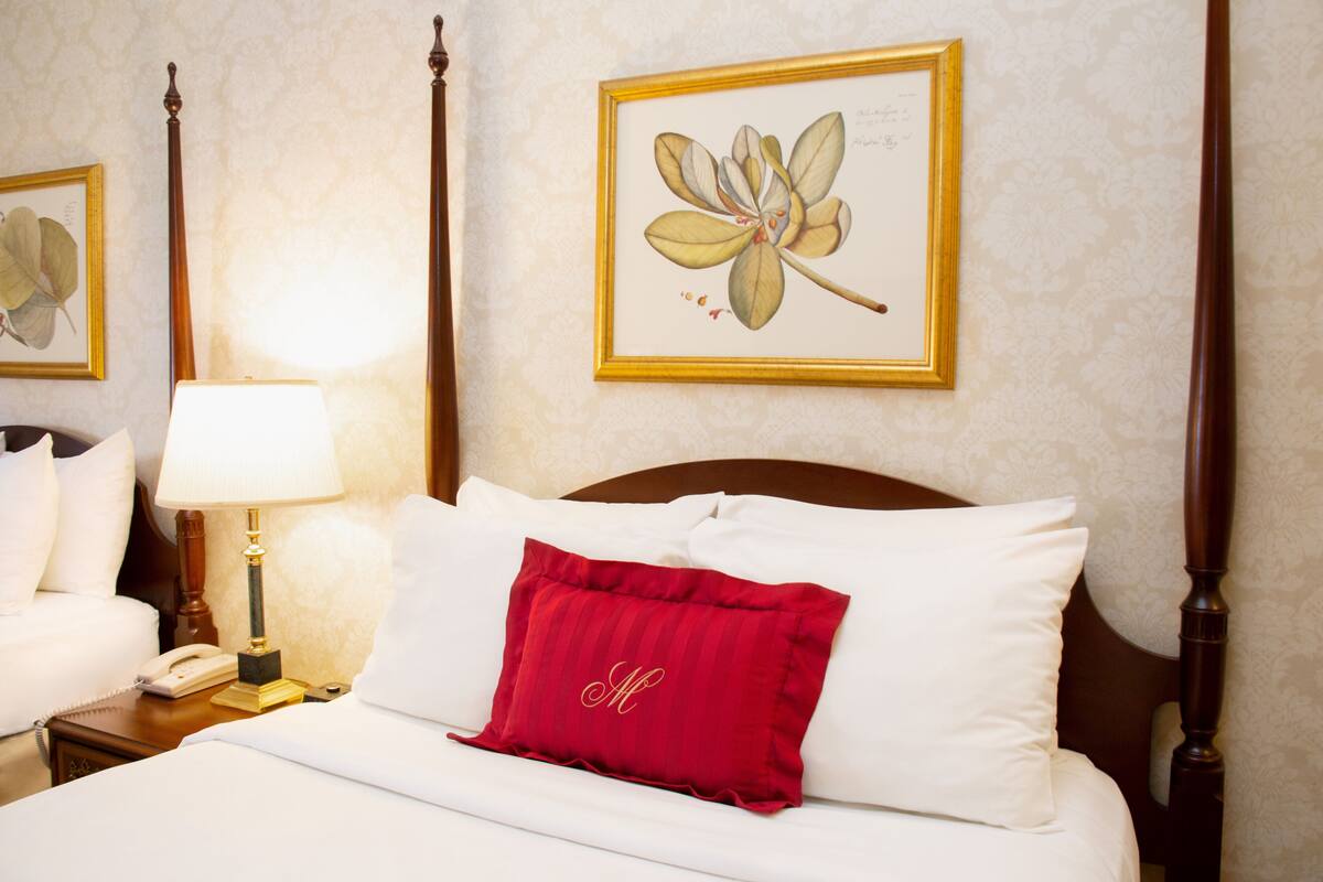 A neatly made bed with white linens and a red pillow, a bedside lamp, a framed botanical print on the wall, and a wooden headboard.