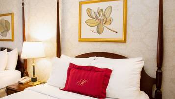 Un lit bien fait, avec du linge de lit blanc et un coussin rouge, une lampe de chevet, une estampe botanique encadrée au mur et une tête de lit en bois.
