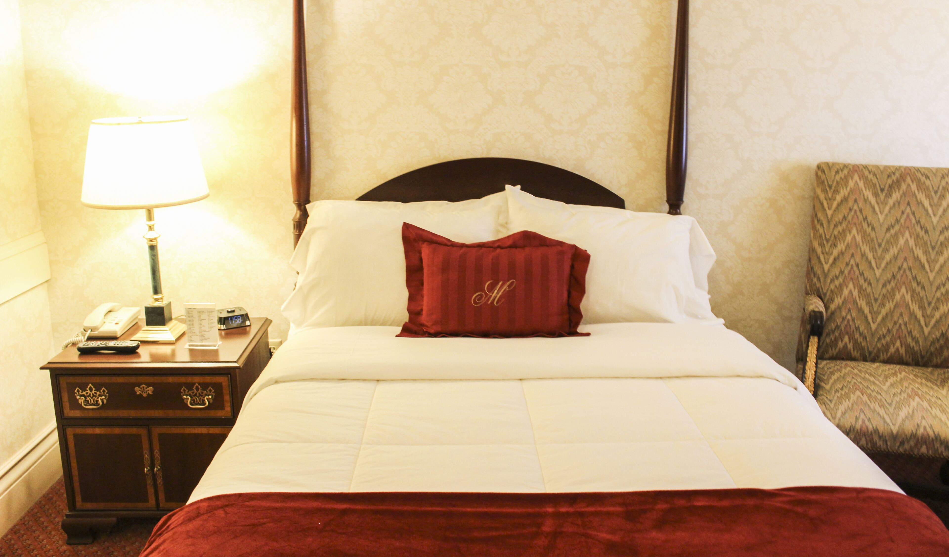 A hotel room with a neatly made bed, a bedside table with a lamp, a telephone, and a clock.