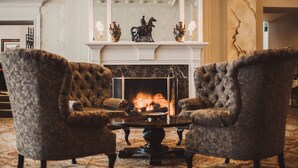 A room with two upholstered armchairs facing a fireplace with a fire, a patterned carpet, and a white mantelpiece with decorative items.