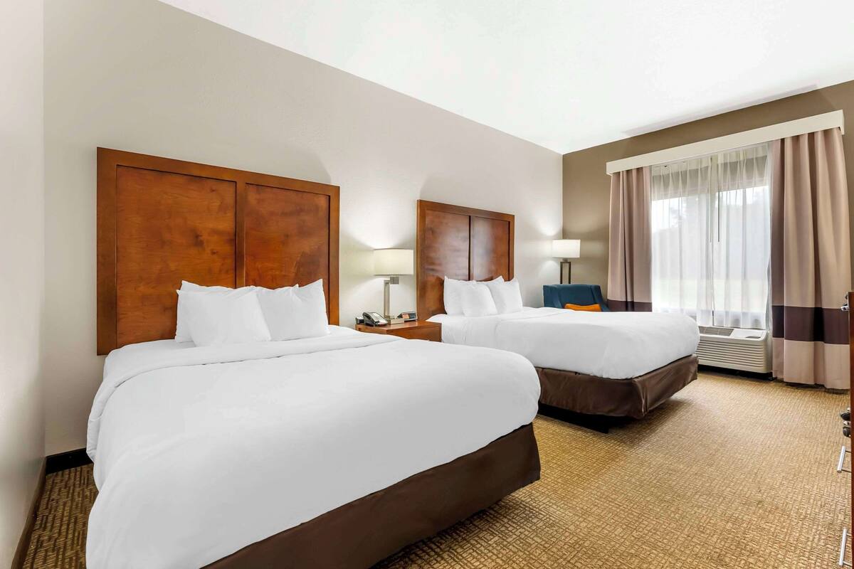 A hotel room with two beds, a nightstand with a lamp, a window with curtains, and a wall-mounted air conditioning unit.