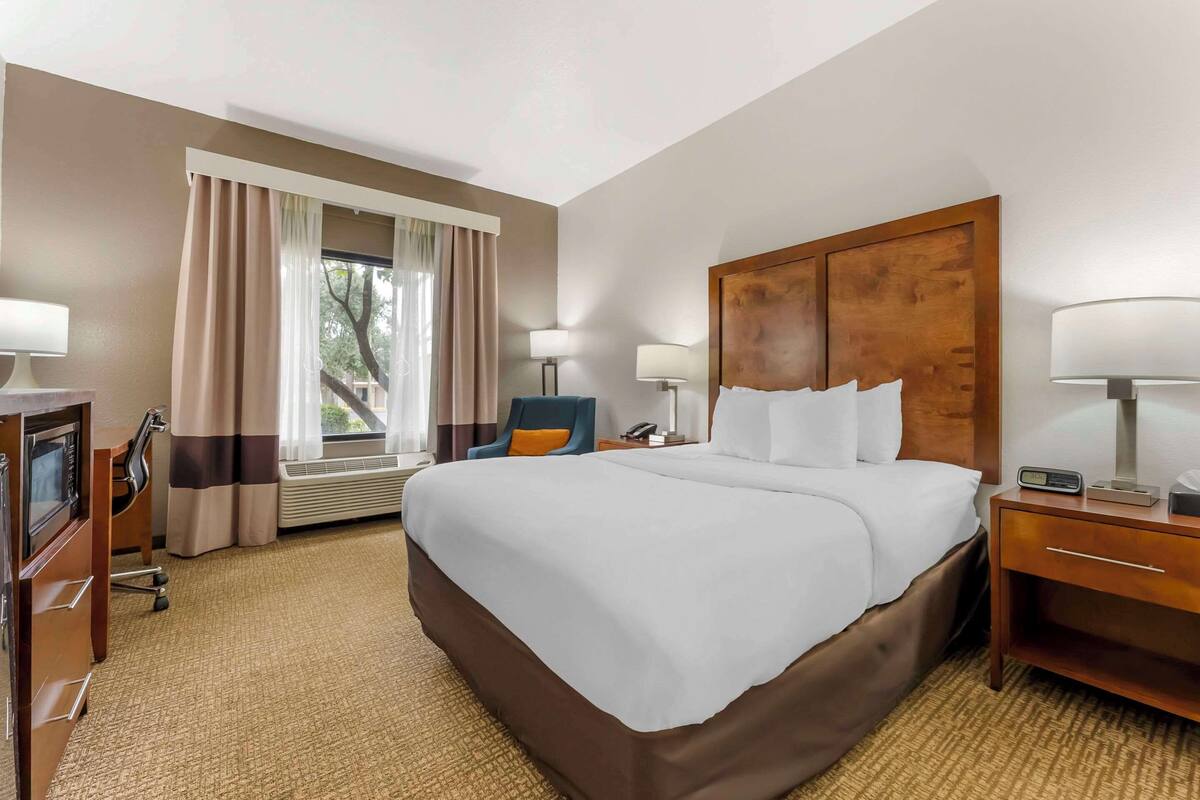 A hotel room with a large bed, a desk with a computer, a chair, a nightstand, and a window with curtains.