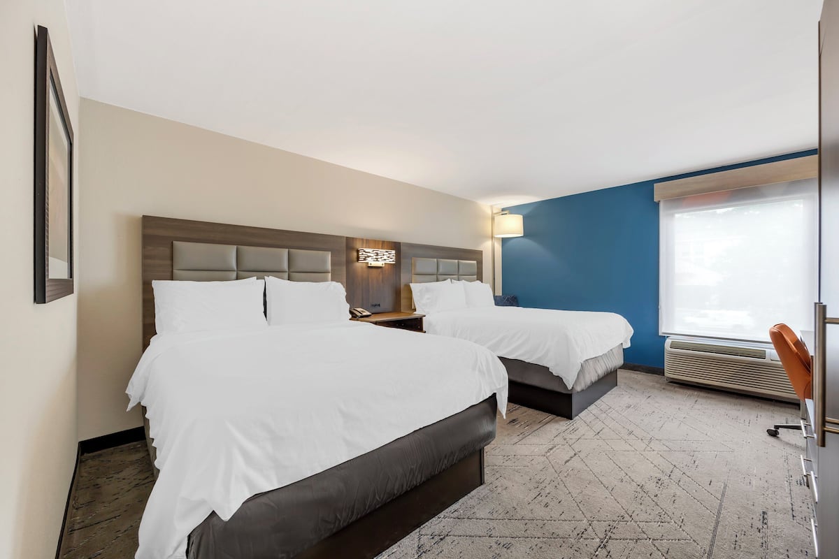 Une chambre d’hôtel avec deux lits, un bureau, une chaise, une fenêtre avec des stores et un climatiseur fixé au mur.