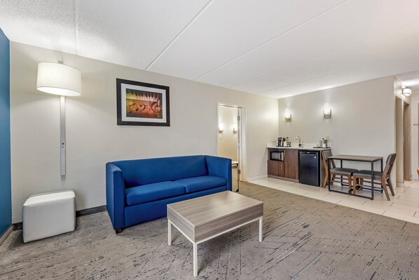 A modern hotel room with a blue sofa, a small white table, a kitchen area with a dining table, and a framed picture on the wall.