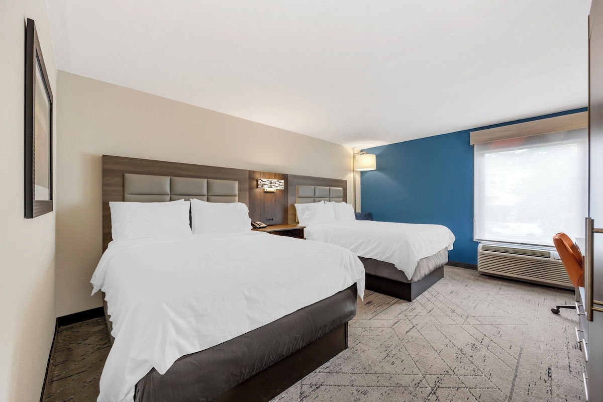 Hotelkamer met twee bedden, een bureau, een stoel, een raam met zonwering en een wandairconditioner.