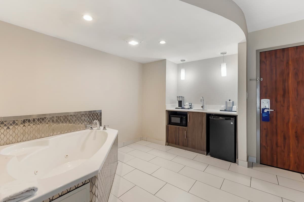 Une salle de bain moderne avec une grande baignoire, un lavabo et un espace douche. On y trouve une porte en bois et un petit coin cuisine équipé d’un four à micro-ondes et d’une machine à café.