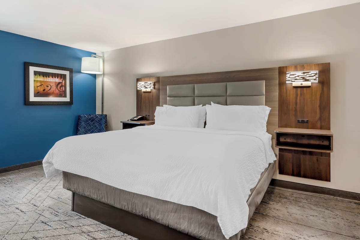 Une chambre d’hôtel avec un grand lit, une table de chevet, une lampe fixée au mur et un tableau encadré accroché au mur.