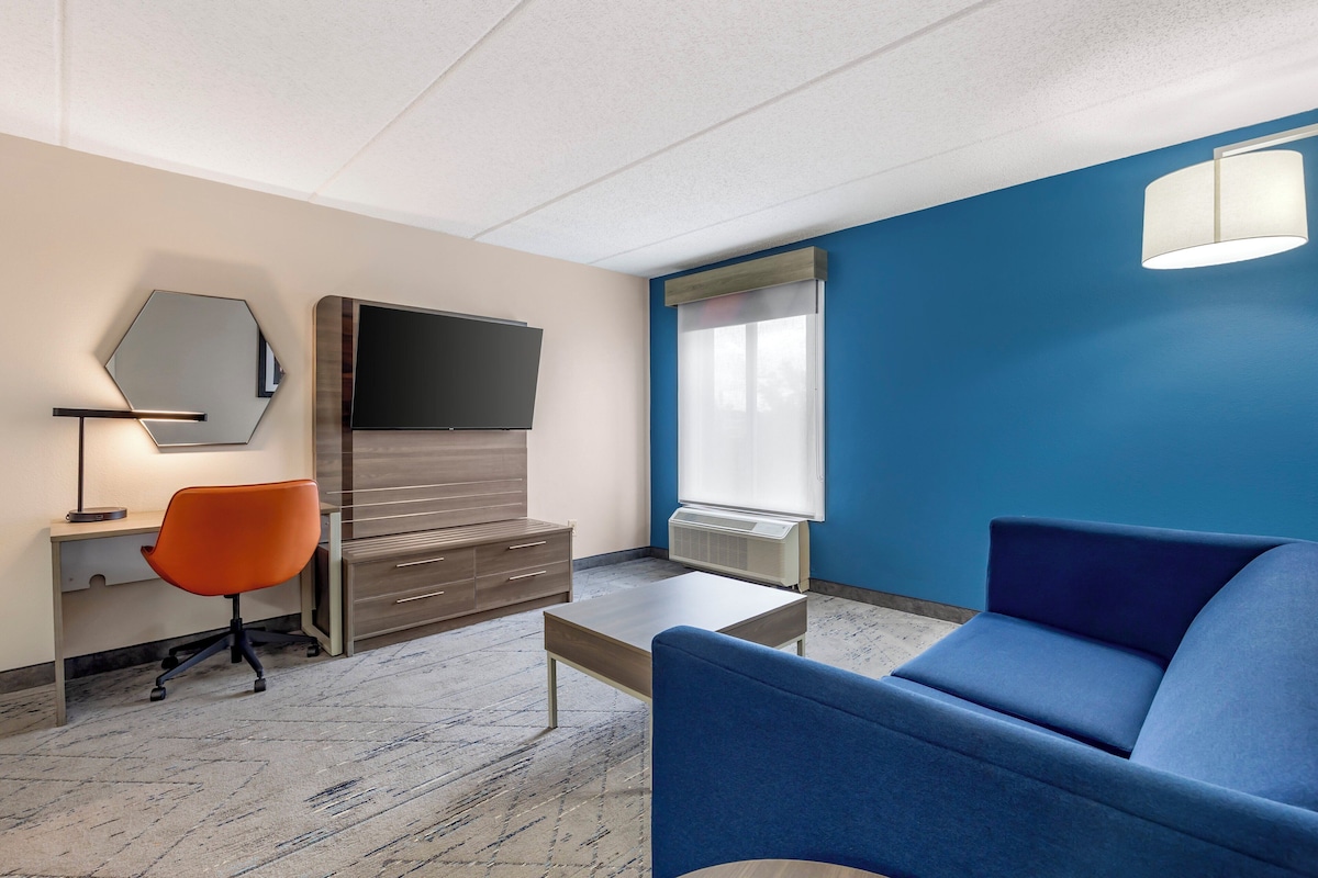 Een moderne hotelkamer met een blauwe bank, een bureau met een oranje stoel, een flatscreen televisie, een grote spiegel en een raam met jaloezieën.