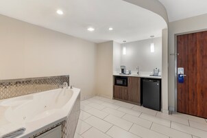 A modern bathroom with a large bathtub, a sink, and a shower area. There is a wooden door and a small kitchen area with a microwave and a coffee maker.