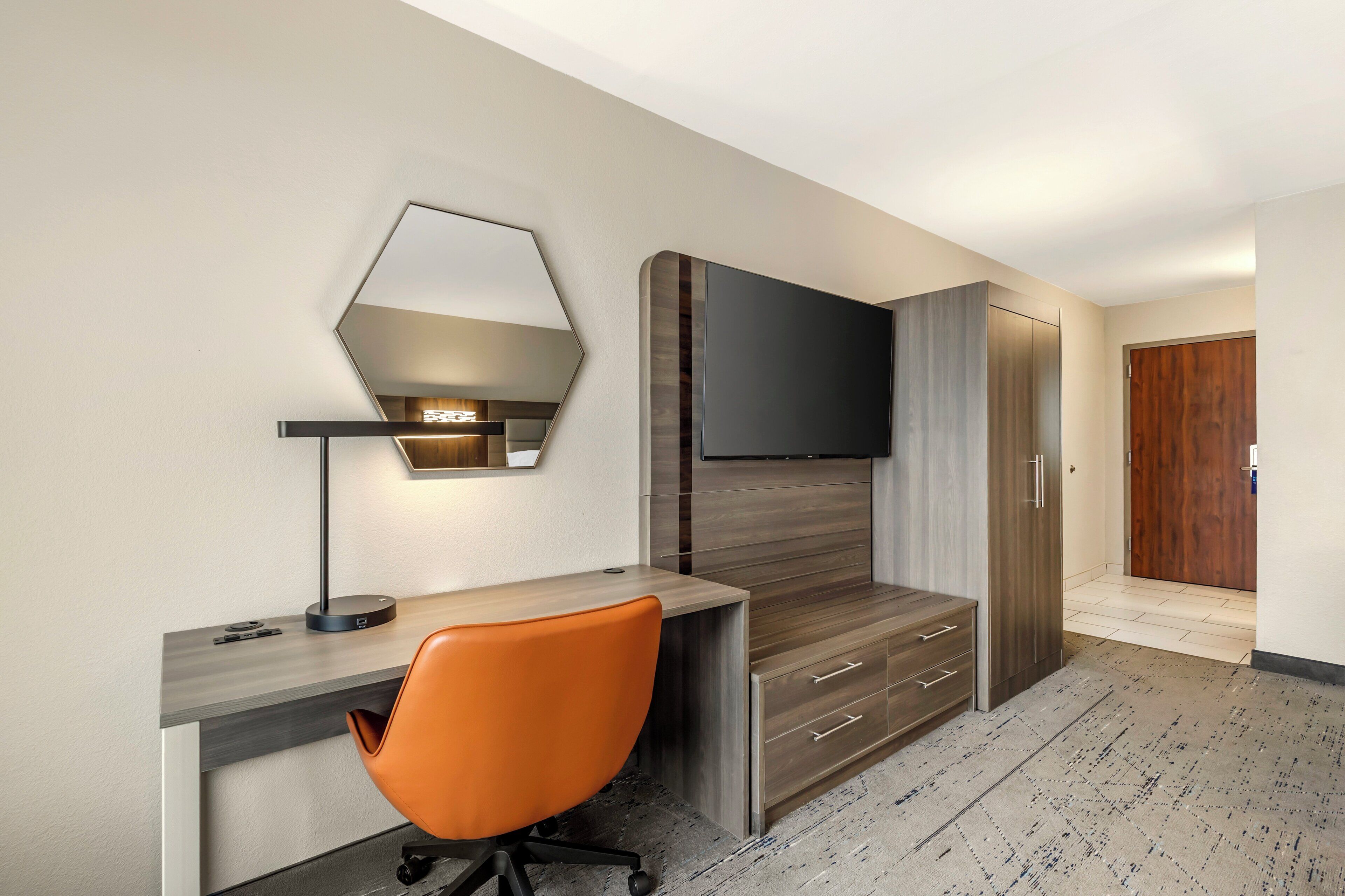 A modern hotel room with a desk, orange office chair, built-in wardrobe, and a flat-screen TV mounted on the wall.