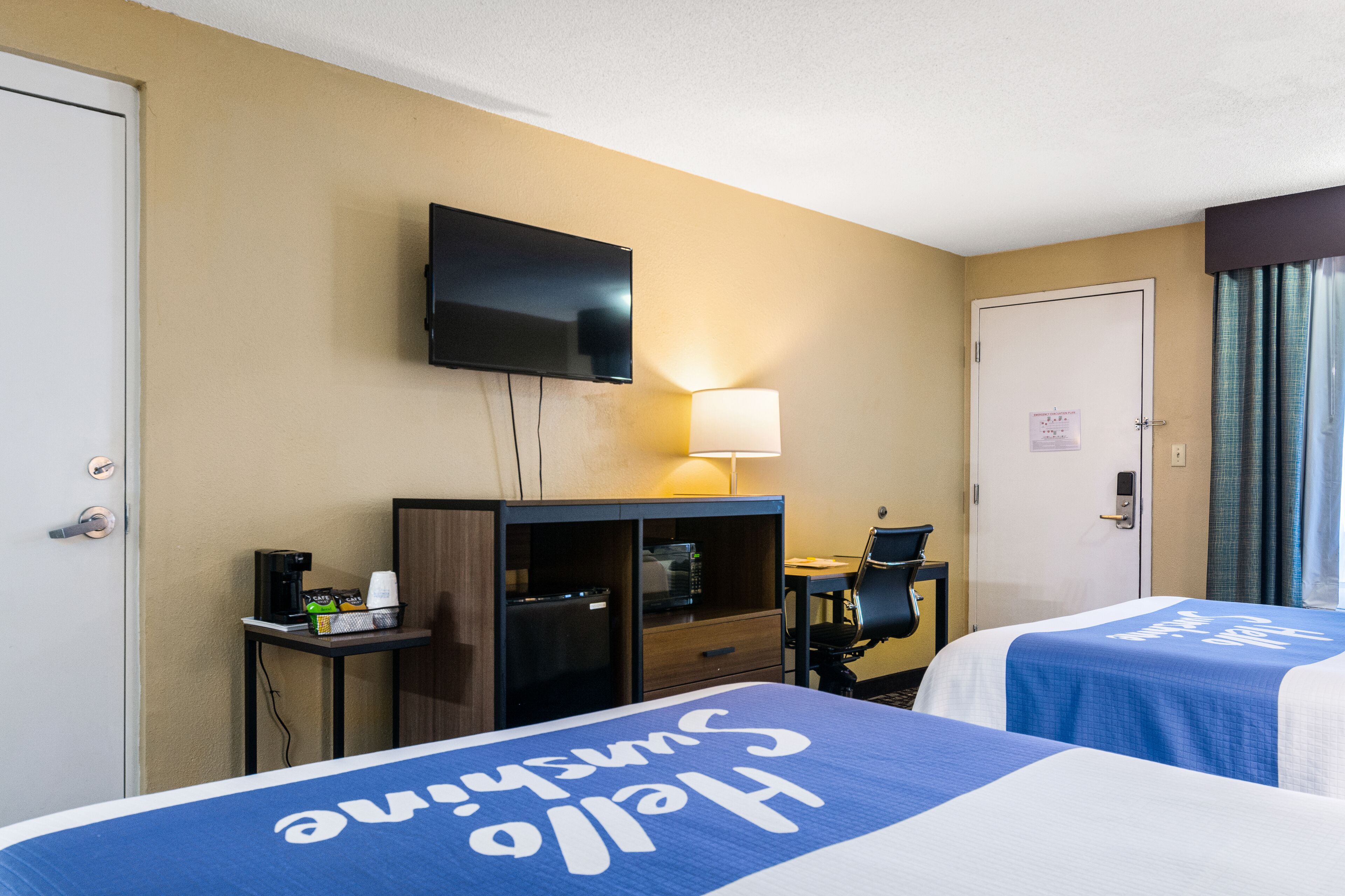 Habitació d'hotel amb dos llits, televisió penjada a la paret, escriptori amb cadira, làmpada i porta amb un cartell.